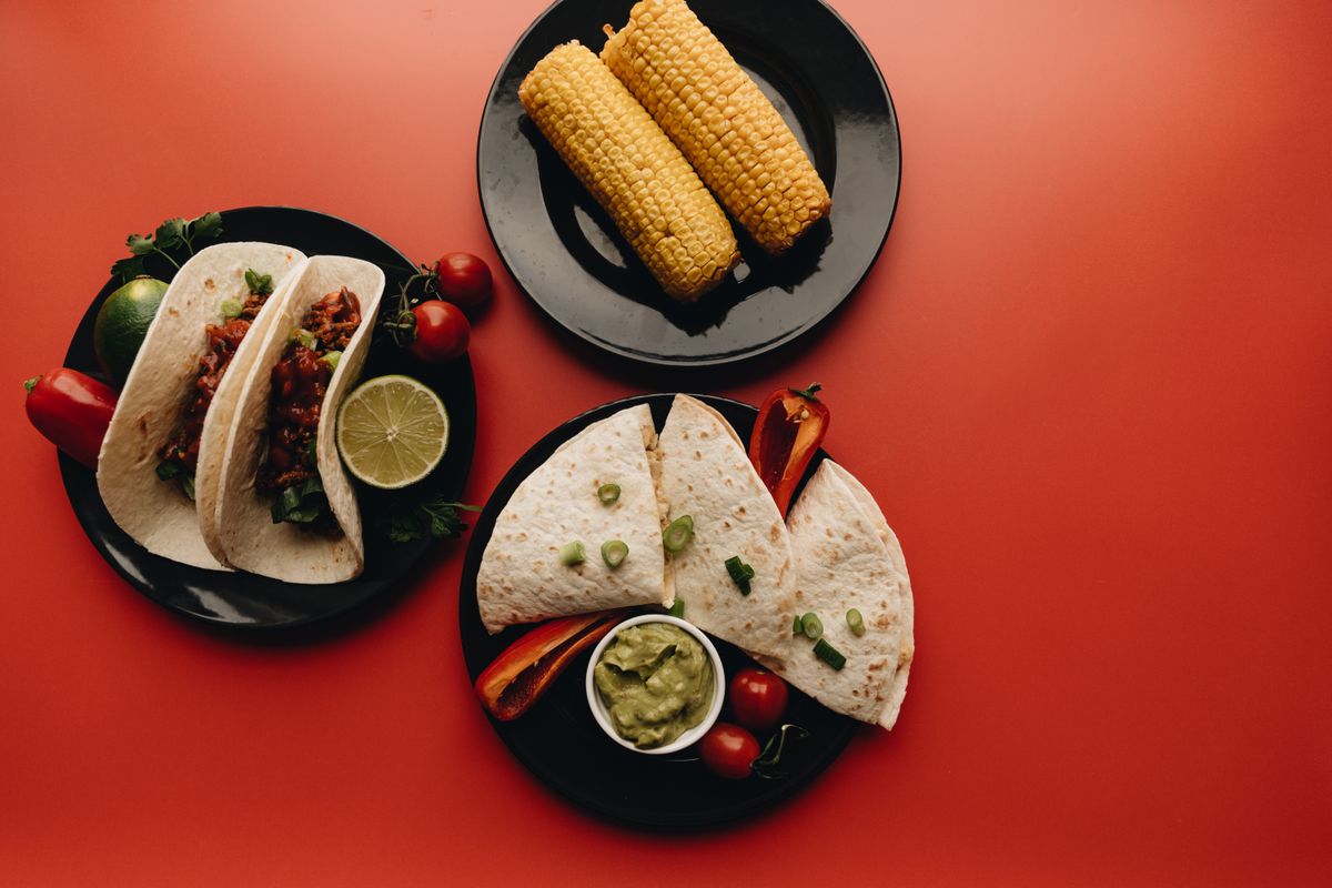 Top view of a spread of Mexican dishes including tacos, corn, and quesadillas on a vibrant red background